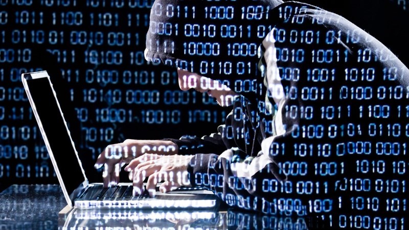 Several rows of binary bits are superimposed over a hooded cyber hacker on a laptop.