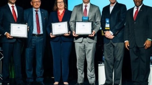 Six business professionals standing on a stage three of them with framed certificates in hand