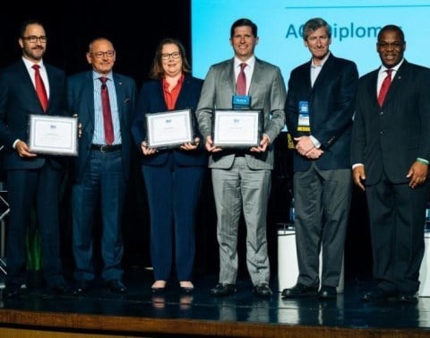 Six business professionals standing on a stage three of them with framed certificates in hand
