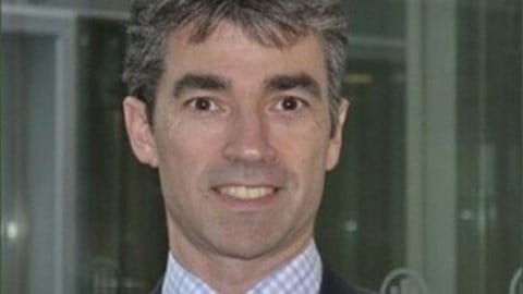 Headshot of Simon Buxton in a grey jacket, plaid shirt, and maroon tie