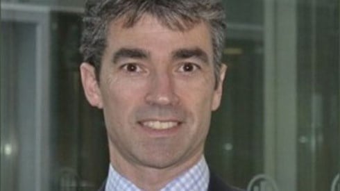 Headshot of Simon Buxton in a grey jacket, plaid shirt, and maroon tie