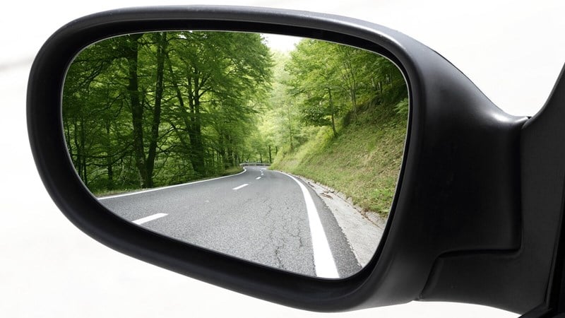 Vechicle rearview mirror showing road with green trees in background