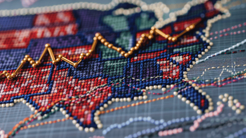 Cross-stitch map of United States with gold pinheads forming an upward-trending line graph