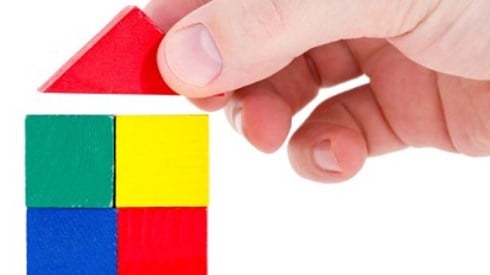 Hand placing triangular red building block on four stacked red, yellow, green, and blue blocks in the shape of a house
