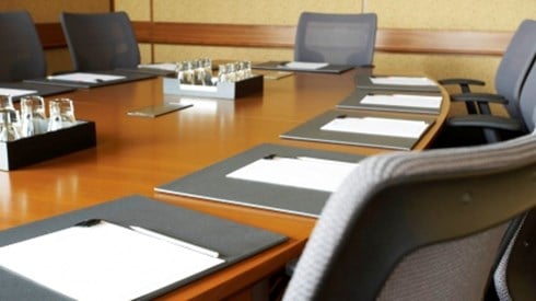 A corporate board room with a long wooden oval table surrounded by gray executive chairs. There are place settings with documents at each chair.