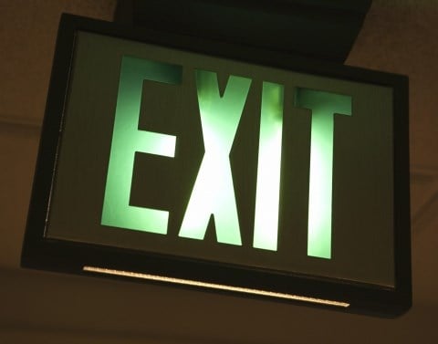A green and white lit up EXIT sign mounted in a black frame on a ceiling