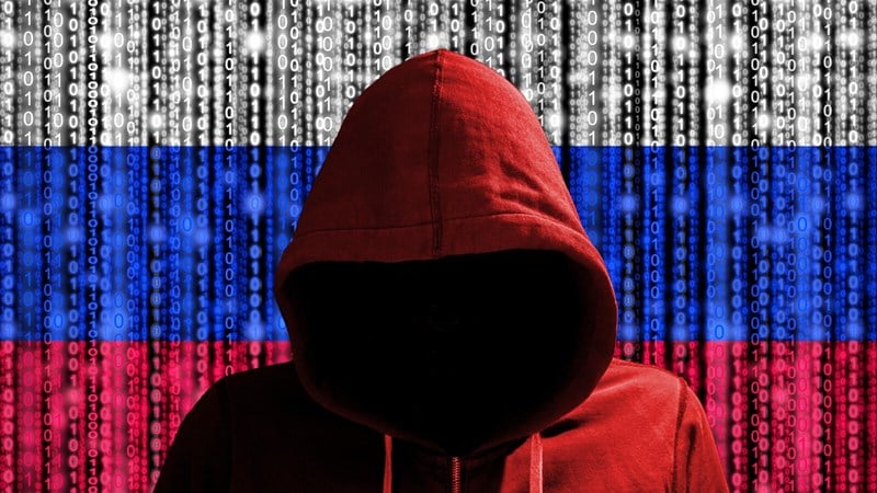 Menacing hacker dressed in a red hoodie standing in front of the Russian Federation flag with computer code on it