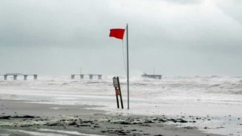 Beach with red warning flag, rough waves, debris, broken pier, and storm clouds