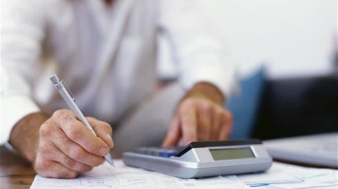 An actuary seated at a desk writing on a paper while using a desk calculator. Documents with charts and diagrams are visible.