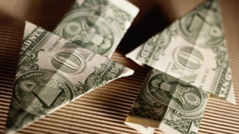 Two one-dollar bills folded in the shape of arrows pointing in opposite directions sitting on brown corrugated cardboard