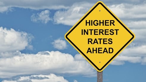 Yellow caution sign reading Higher Interest Rates Ahead against blue sky with clouds