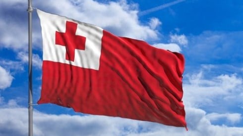 Against a blue sky flies the flag of Tonga which is red and in the left corner has a white square with a red cross