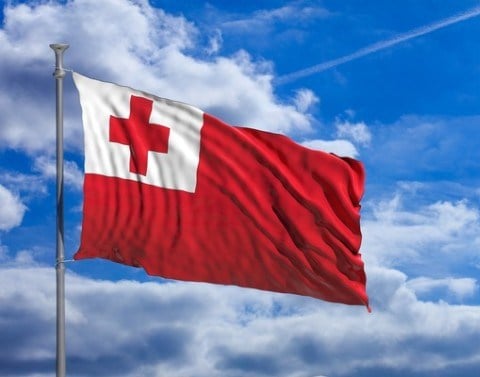 Against a blue sky flies the flag of Tonga which is red and in the left corner has a white square with a red cross