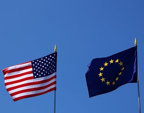 United States and European Union flags flying on poles with blue sky background