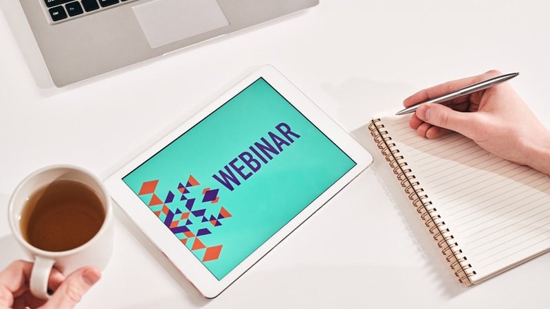 Tablet showing the word Webinar with a computer and pen and paper nearby