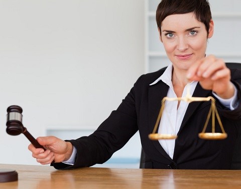 Woman judge holding a gavel in one hand and gold colored scales of justice in the other hand while sitting at a wooden desk