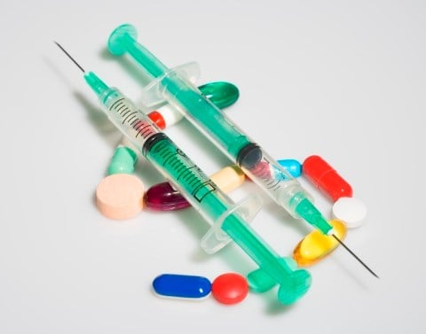 Two syringes with needles resting on top of pills forming the shape of a dollar sign