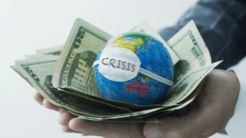 Hand holding globe with face mask on it on top of pile of dollar bills