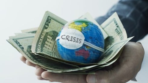 Hand holding globe with face mask on it on top of pile of dollar bills