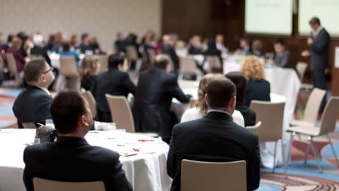 Business professionals seated at round tables in a conference hall listen to a speaker give a presentation.
