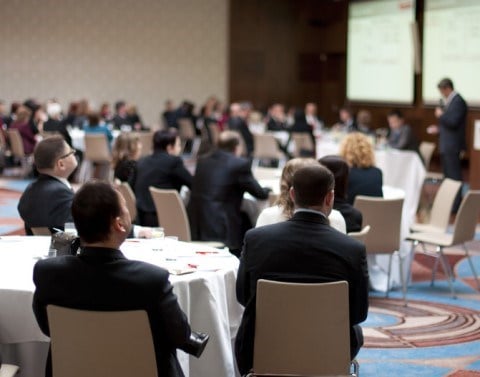 Business professionals seated at round tables in a conference hall listen to a speaker give a presentation.