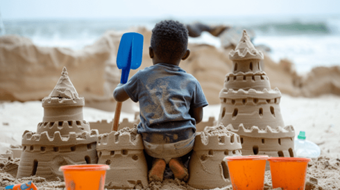 A toddler building a sand castle on the beach and holding a blue plastic shovel in his left hand with three orange plastic buckets next to him