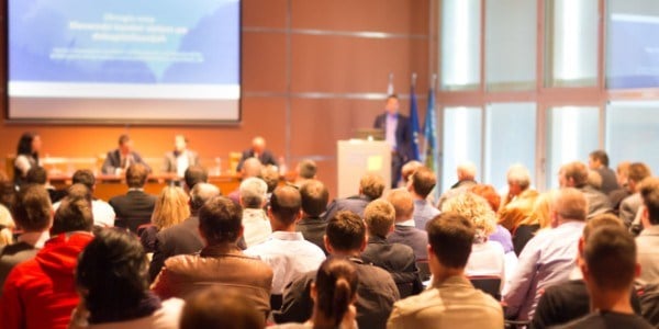 Group of people viewing a slide presentation in a large room with speaker at podium, panel of experts, and 3 flags in corner
