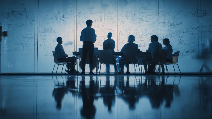 group of businesspeople sitting around a conference table in a blue room, one person standing and writing on the wall