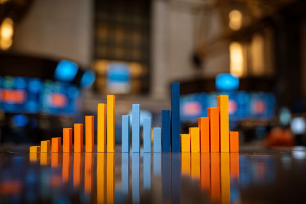 blue and orange bar chart of varying heights and its reflection in front of a blurred stock exchange room
