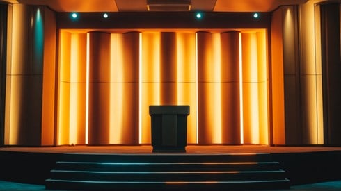 An empty conference stage lit with golden light, and a few stairs leading up to a podium in the center of the stage.