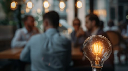 An illuminated light bulb resting on a wooden table in the foreground, with a blurred background of businesspeople in a conversation.
