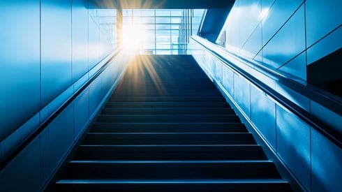 A staircase in a corporate office, leading upwards to bright sunlight shining through the lobby windows.