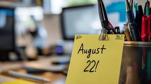 Close-up of a Post-It note on a cluttered desk with "August 22!" written on the note