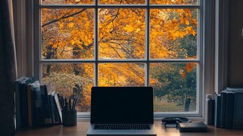 A desk with a laptop on it in front of a window with tree leaves turning yellow