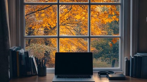 A desk with a laptop on it in front of a window with tree leaves turning yellow