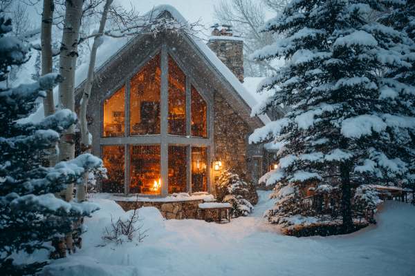 cozy winter scene of a house in a wooded area covered in snow and snowflakes showing a warm, amber, glowing fireplace through a large picture window