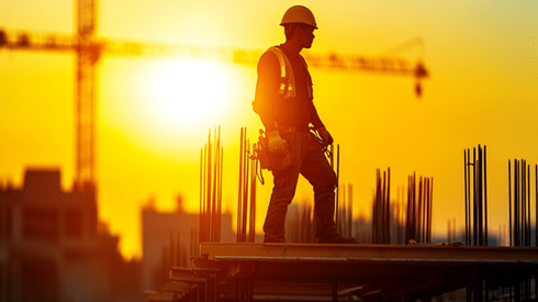 A construction worker on the roof of a jobsite is silhouetted again a strong setting sun with a crane in the background