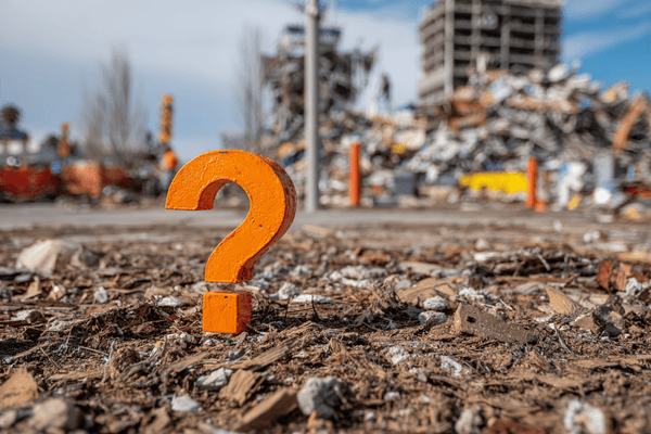 An orange question mark in the foreground with damaged buildings in the background