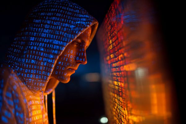 hooded hacker sitting near a screen with projected orange and blue binary code onto him