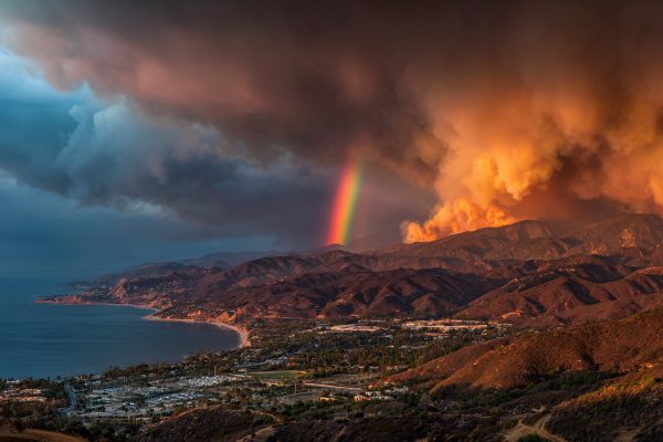 storm clouds and wildfire smoke over a coastal city with a rainbow shining down behind a mountain