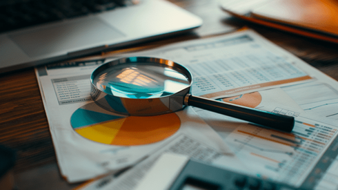 A magnifying glass sits on top of papers with tables, pie charts, and graphs next to a calculator and laptop