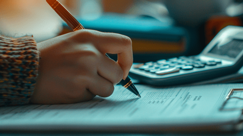 A hand is holding a pen to papers on a clipboard with a calculator sitting nearby