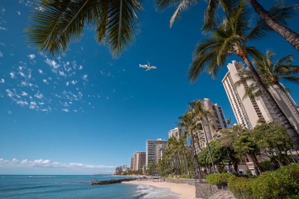 Hawaiian beach with commercial buildings and palm trees and a plane flying in overhead
