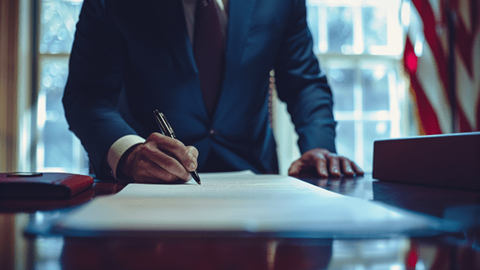 A man in a suit signs a piece of paper laying on his desk with an American flag in the background