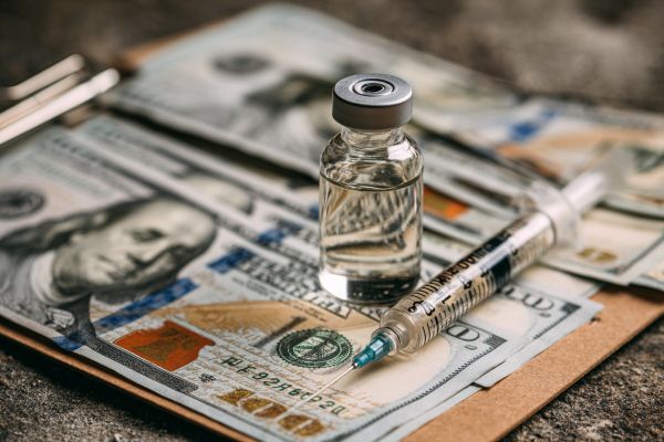 clear medication vial and a syringe with a needle on a clipboard and hundred dollar bills