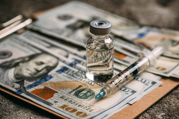 clear medication vial and a syringe with a needle on a clipboard and hundred dollar bills