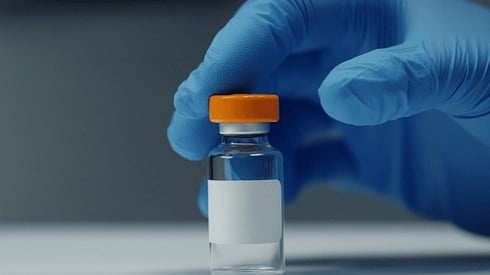A hand in a blue latex glove reaches for a glass vaccine bottle with an orange lid