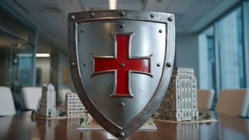 silver knight's shield with a red cross in front of a model commercial business building on a conference table