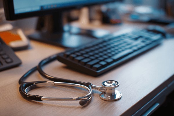 A stethoscope on an office desk next to a computer keyboard and monitor