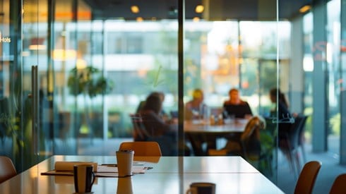 A conference table with coffee mugs and empty chairs in the foreground, while a small business team meets in a separate room with full glass windows in the background.